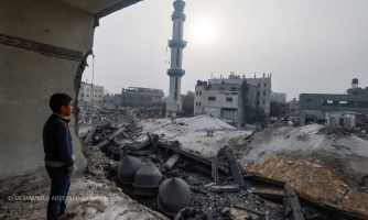 Lapsi seisoo palasiksi pommitettujen harmaiden rakennusten edustalla ja katselee niitä.