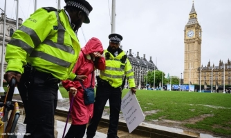 Kaksi poliisia taluttaa iäkästä ihmistä, jolla on punainen takki ja kävelykeppi. Taustalla Big Ben.