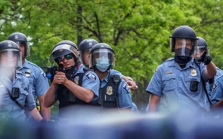 Poliisien joukko, yksi heistä osoittamassa joukkojenhallinta-aseella kuvaajaan päin.