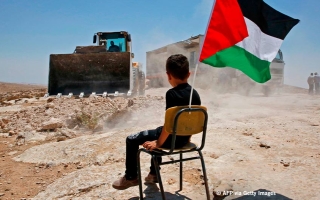 Lapsi istuu aavikolla tuolilla, johon on kiinnitetty Palestiinan lippu, ja katsoo suurta kaivinkonetta.