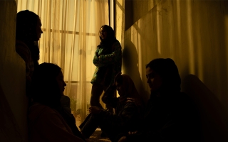 Viisi naista huivit päässä ja kasvot varjossa huoneessa ikkunan takana.