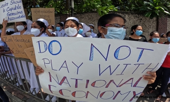 Mielenosoitus. Etualalla on kasvomaskiin pukeutunut ihminen, joka pitää kädessään kylttiä, jossa lukee: "Do not play with national economy)