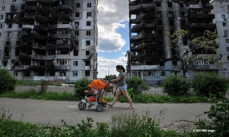 T-paitaan ja farkkusortseihin pukeutunut ihminen työntää oransseja lastenvaunuja pommituksissa tuhoutuneiden rakennusten edessä Ukrainassa.
