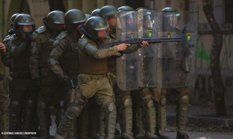 Vihreisiin uniformuihin ja kypäriin pukeutuneet sotilaat seisovat rivissä. Osalla on joukkojenhallintakilpiä. Etualalla yksi henkilö osoittaa eteensä kiväärillä.