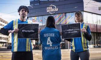 Kolme aktivistia Fifa-museon edustalla. Kaksi aktivistia pitää kylttejä, joissa lukee #PayUpFIFA. Kolmas ihminen on selin kameraan ja hänen paidassaan lukee Justice 4 workers.