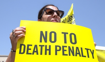 Aurinkolaseihin pukeutunut ihminen pitää käsissään keltaista mielenosoituskylttiä, jossa lukee mustilla kirjaimilla "no to death penalty".