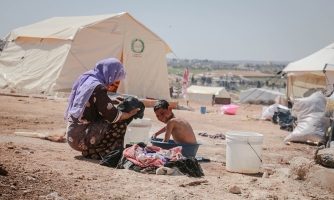 Nainen ja poika pakolaisleirillä. Nainen on kyykistynyt pienessä vadissa peseytyvän pojan eteen, poika katsoo hymyillen kameraa.